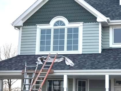 House exterior with ladder, green siding, large windows, and gray roof.