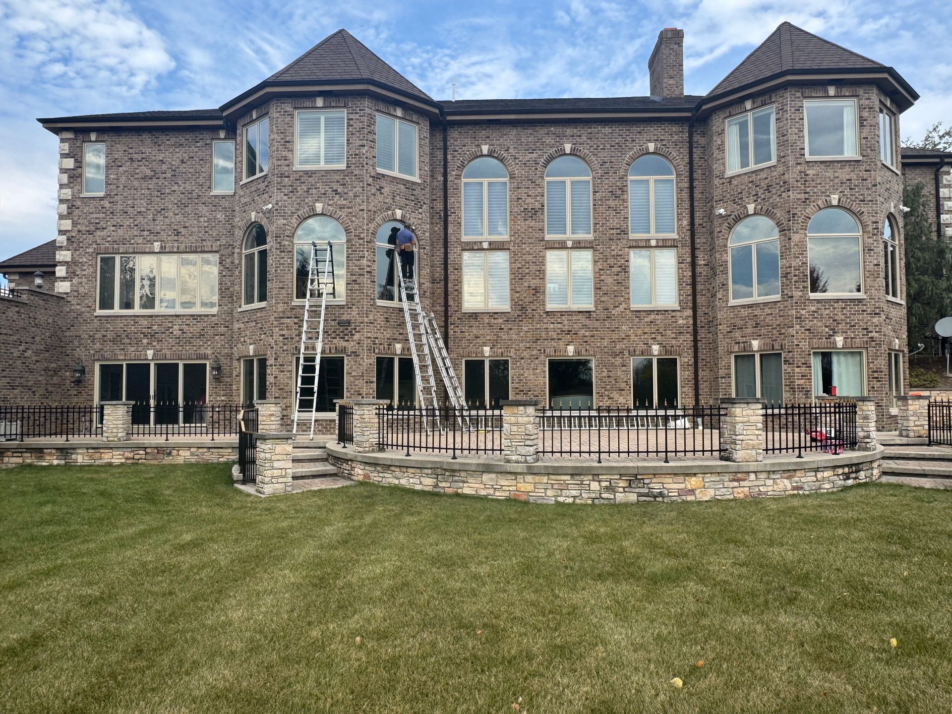 Large brick house with multiple windows, two people on ladders, and a green lawn.