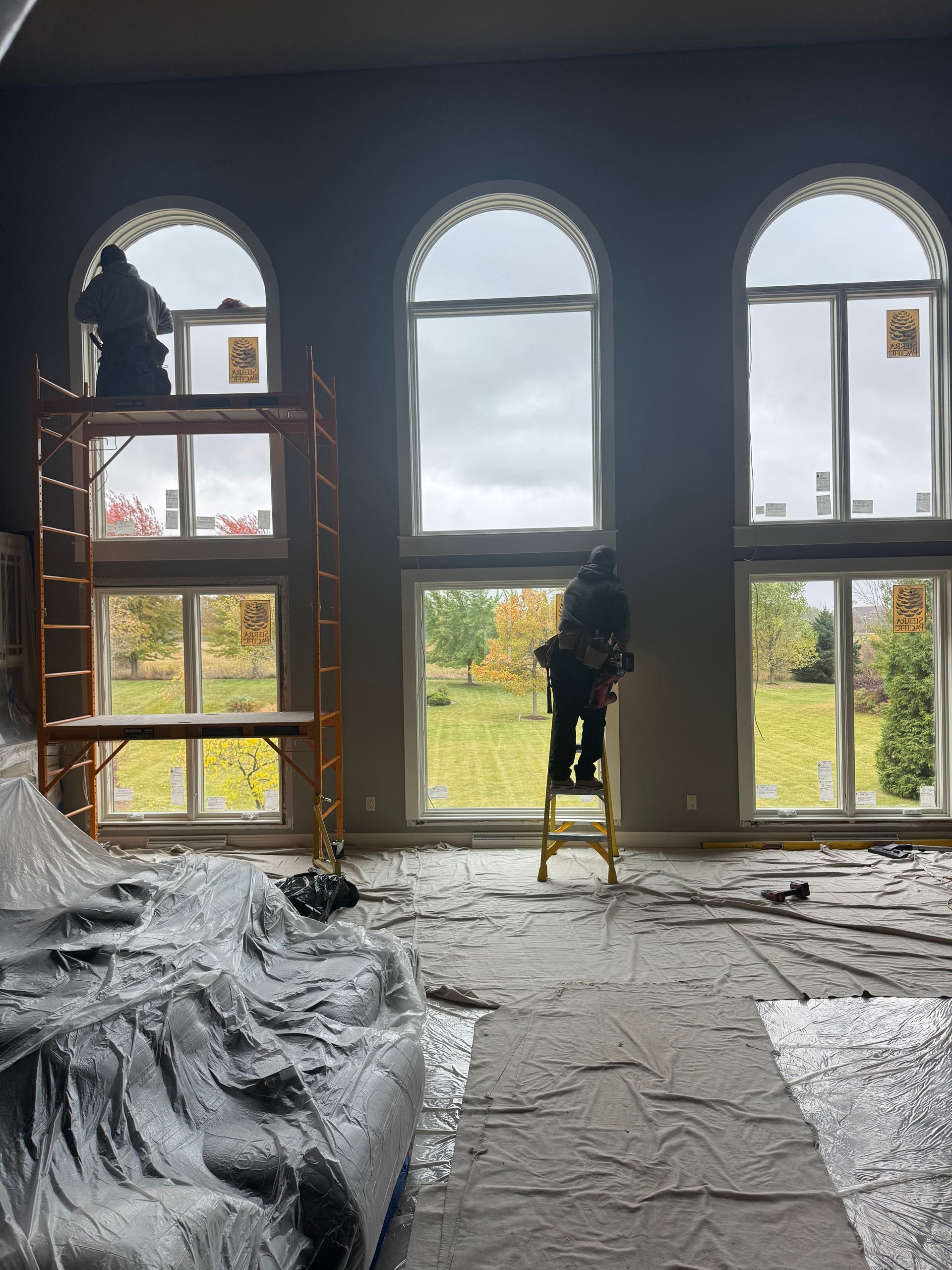 Two workers installing windows in a large room with arched windows; one on a scaffold, the other on a ladder.