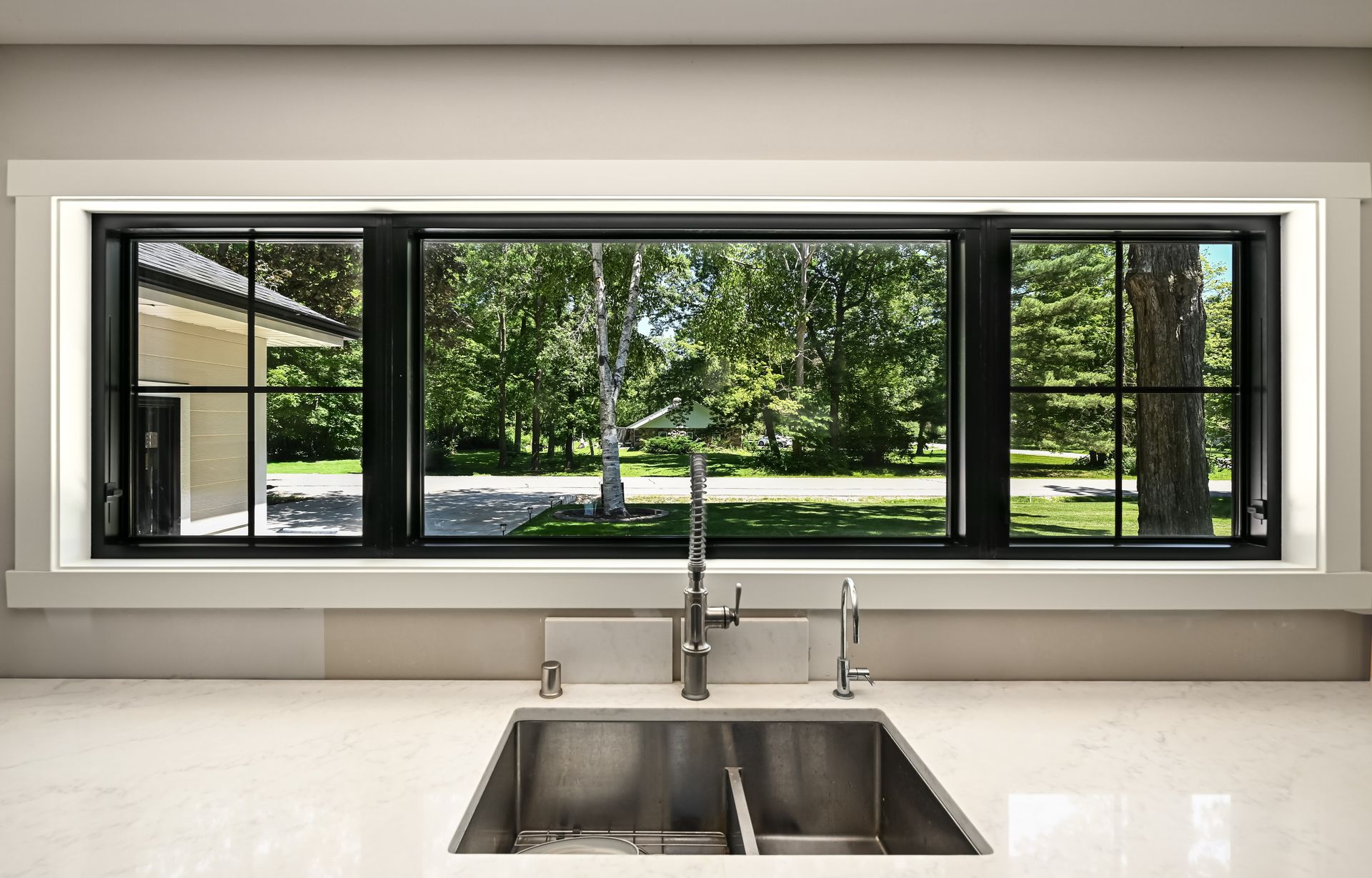 Kitchen window overlooking trees and yard, with black frame, stainless steel sink and faucet.