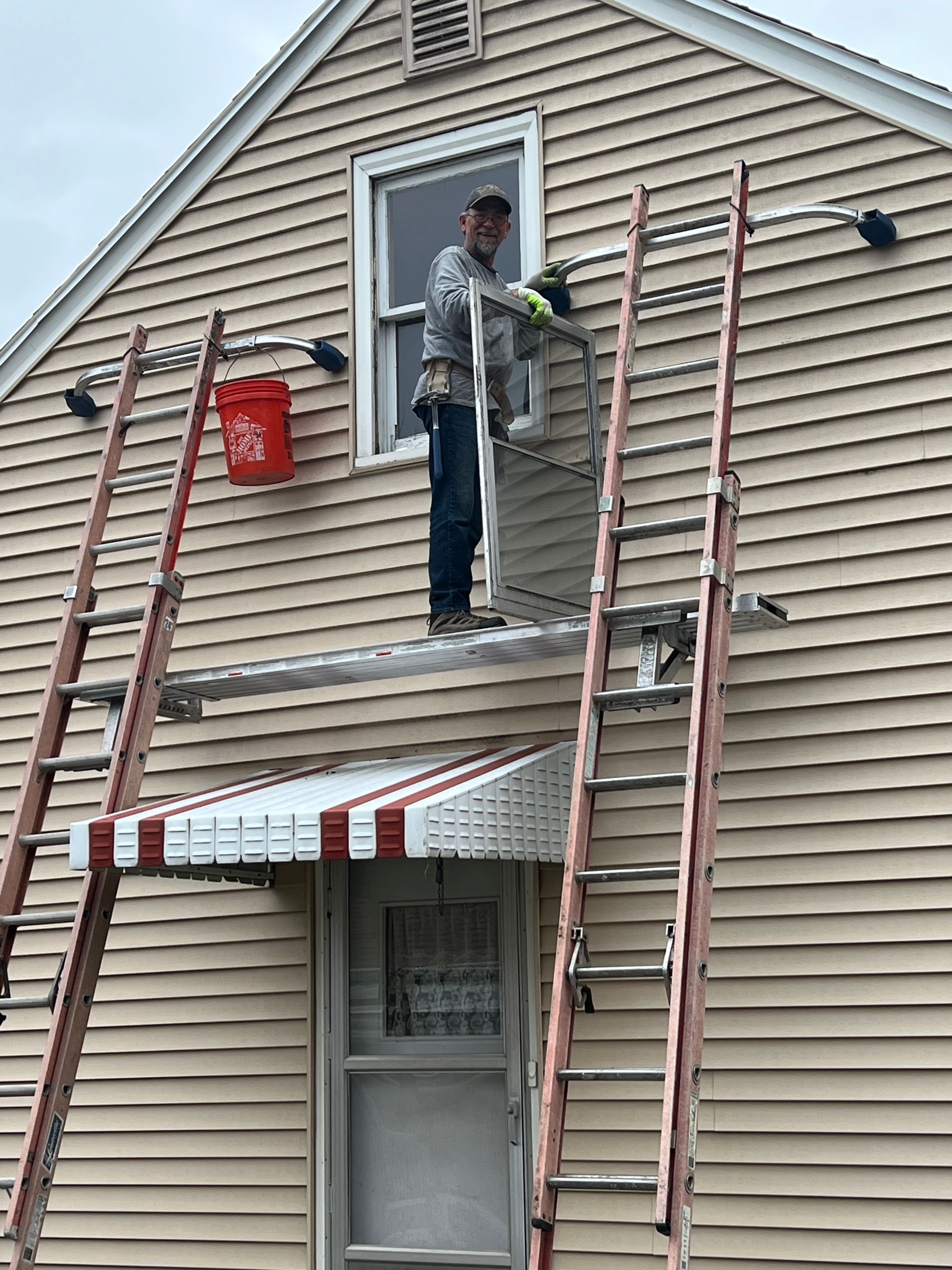 A person standing on scaffolding, working on a window on a house. Red bucket, two ladders, and awning below.