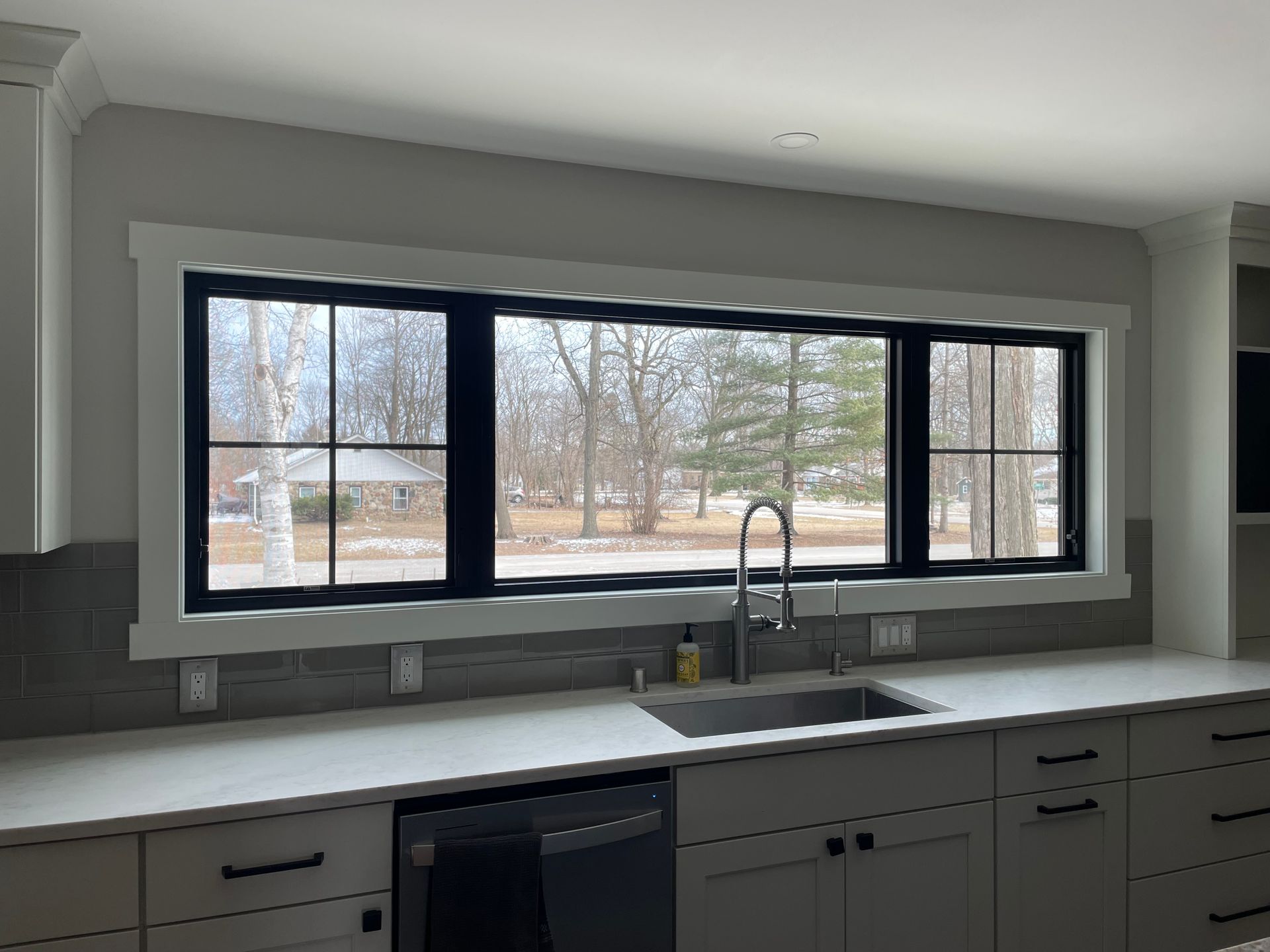 Kitchen with black-framed windows above a countertop and sink, looking out at trees.