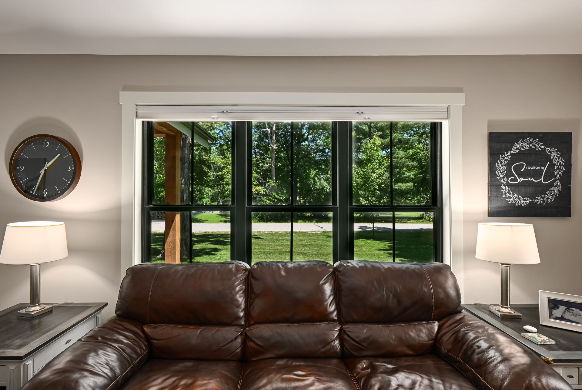 Brown leather sofa in front of a window overlooking greenery. Side tables with lamps flank it.