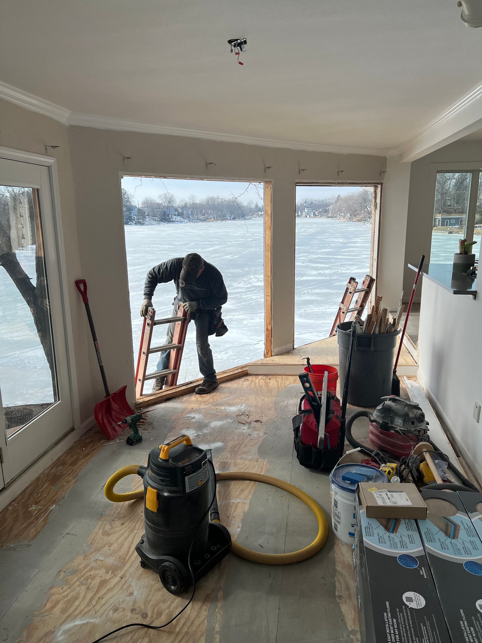 Person on a ladder near a large window, working inside a room with construction tools. Outside, a frozen body of water.