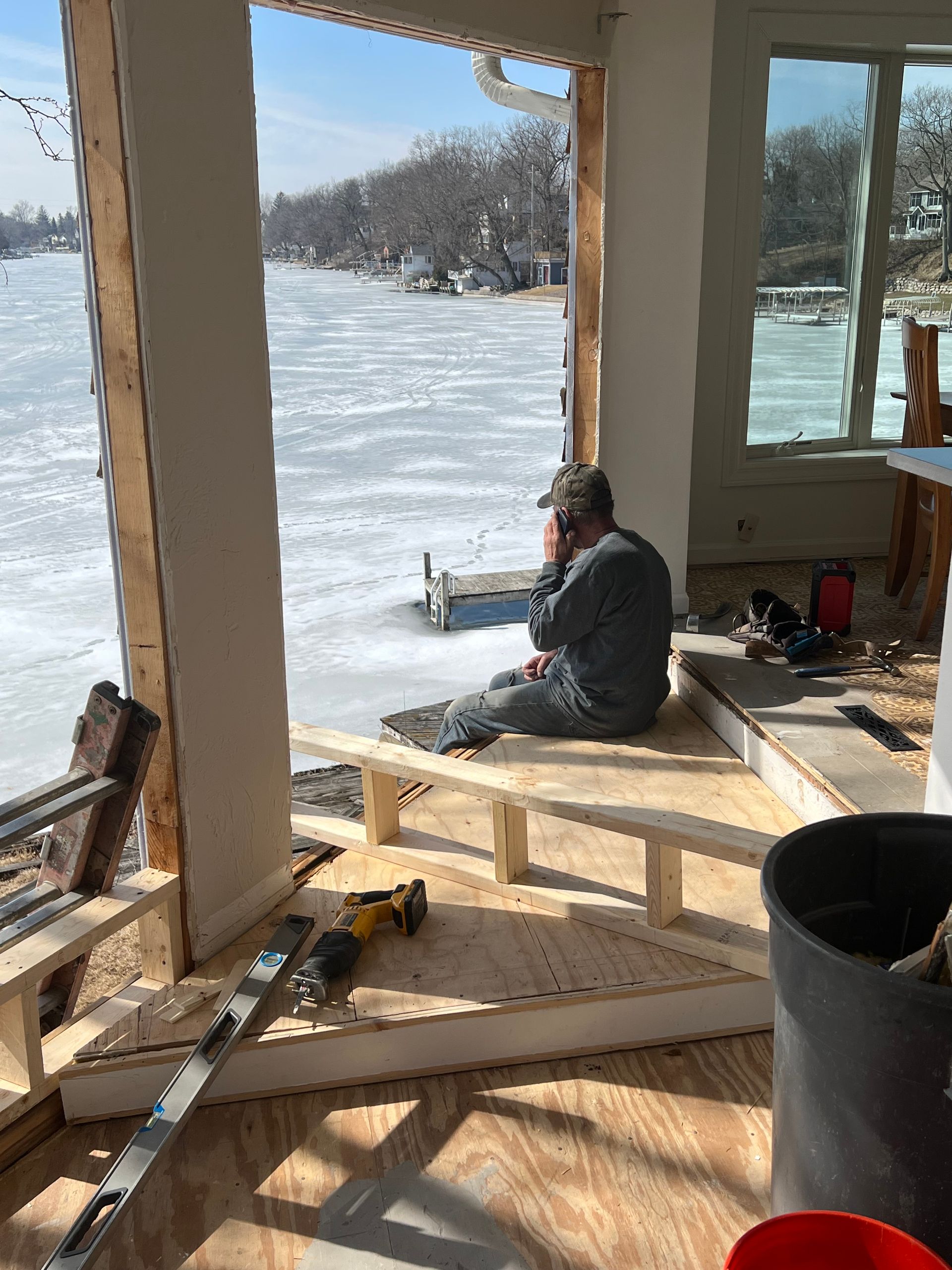Man sits on a platform inside a sunroom, looking out at a frozen lake. Construction tools are nearby.