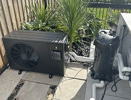 Black pool heater and filter system on a patio, next to greenery.