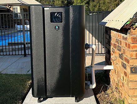 Black pool heater next to a brick wall and metal fence, blue pool in the background.