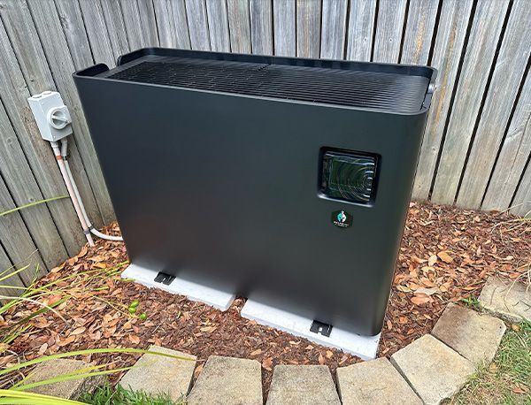 Black pool heater with digital display outdoors, on a gravel bed, next to a wooden fence.