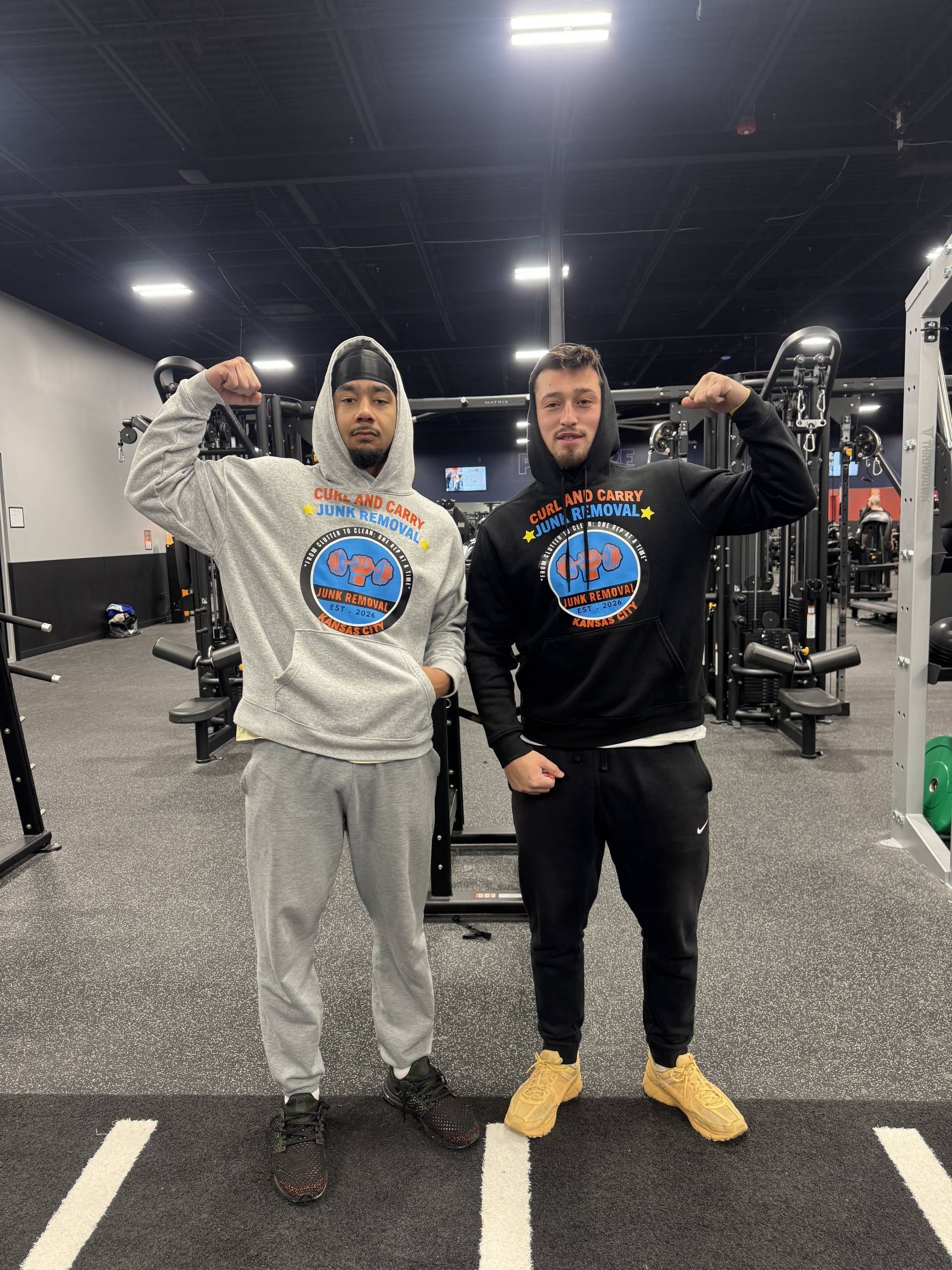 Two men in matching hoodies pose in a gym, each flexing one arm toward the camera.