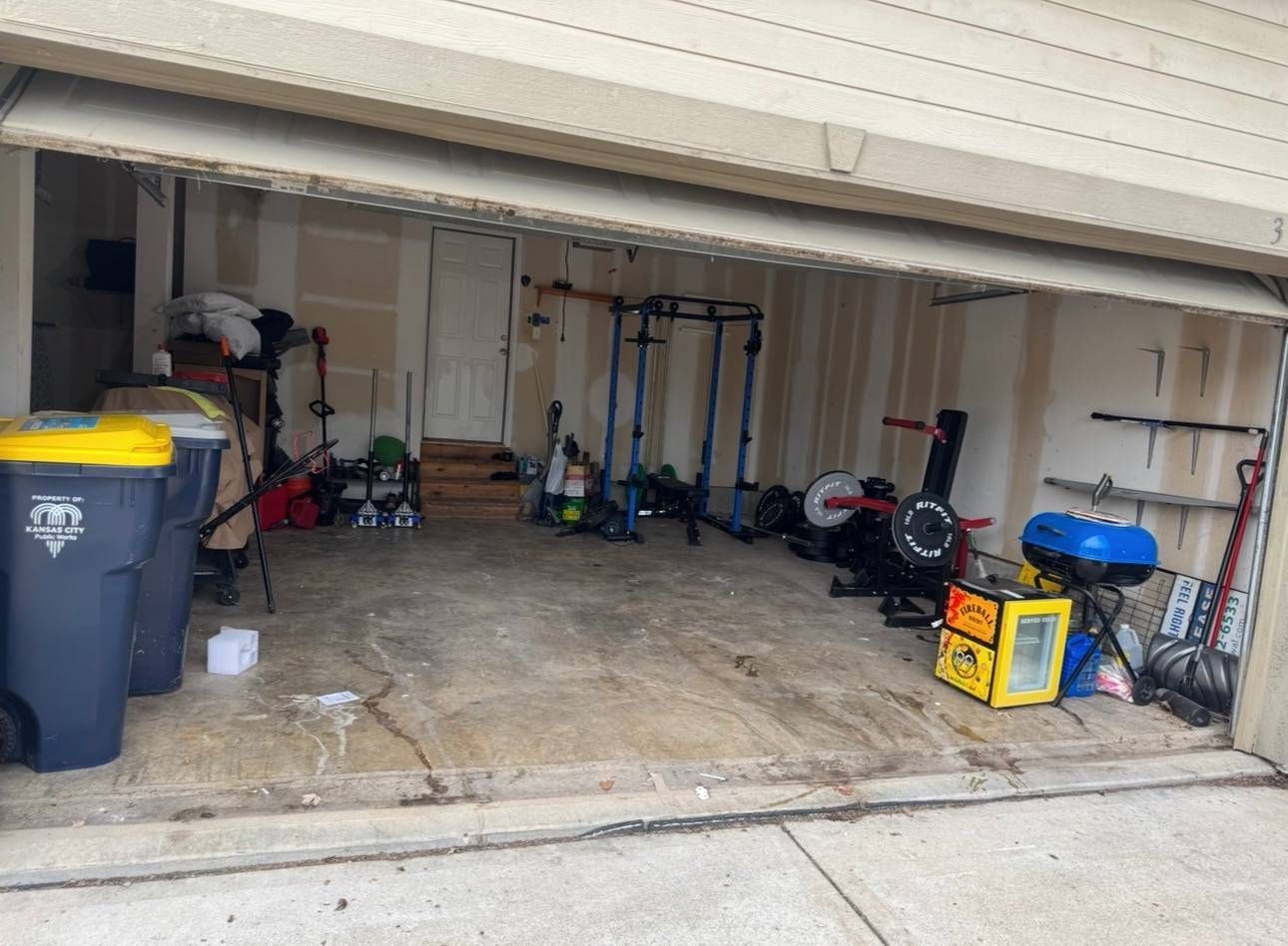 Open residential garage containing a power rack, fitness equipment, a yellow cooler, and large trash bins.