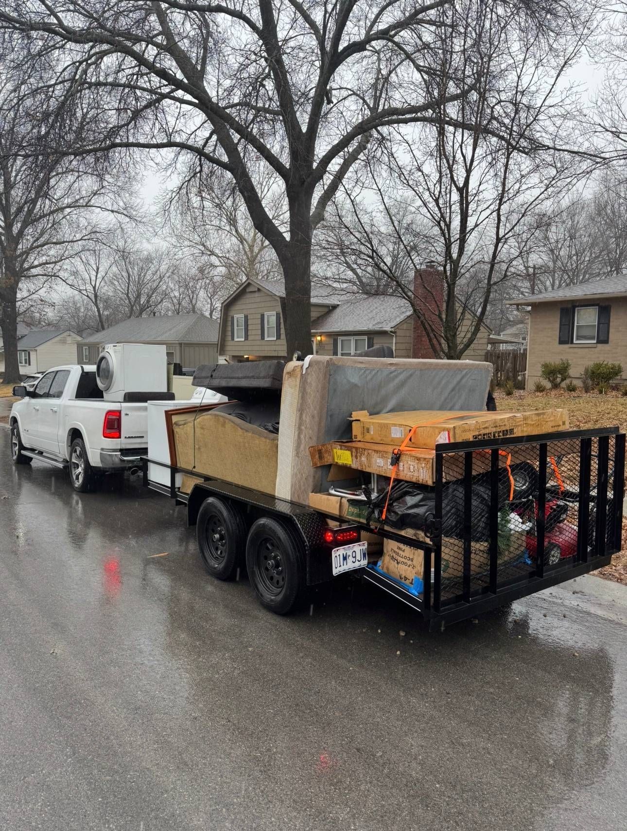 A white pickup truck hauls a loaded utility trailer filled with a mattress, furniture, and boxes on a rainy street.