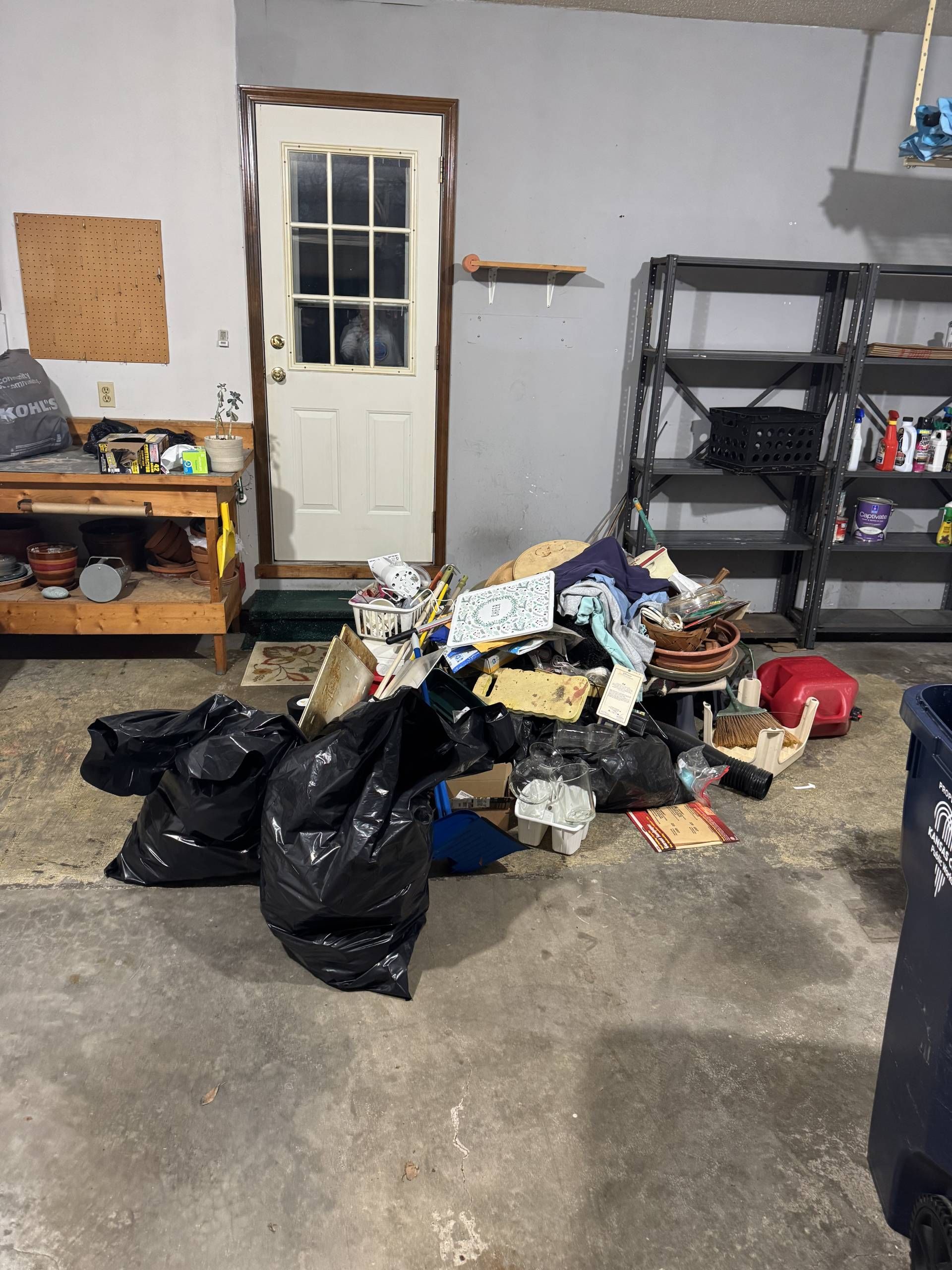 A cluttered garage with black trash bags, cardboard boxes, and miscellaneous items piled on the concrete floor.