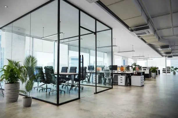 A modern, open-plan office featuring a glass-walled conference room with black metal frames and potted green plants.