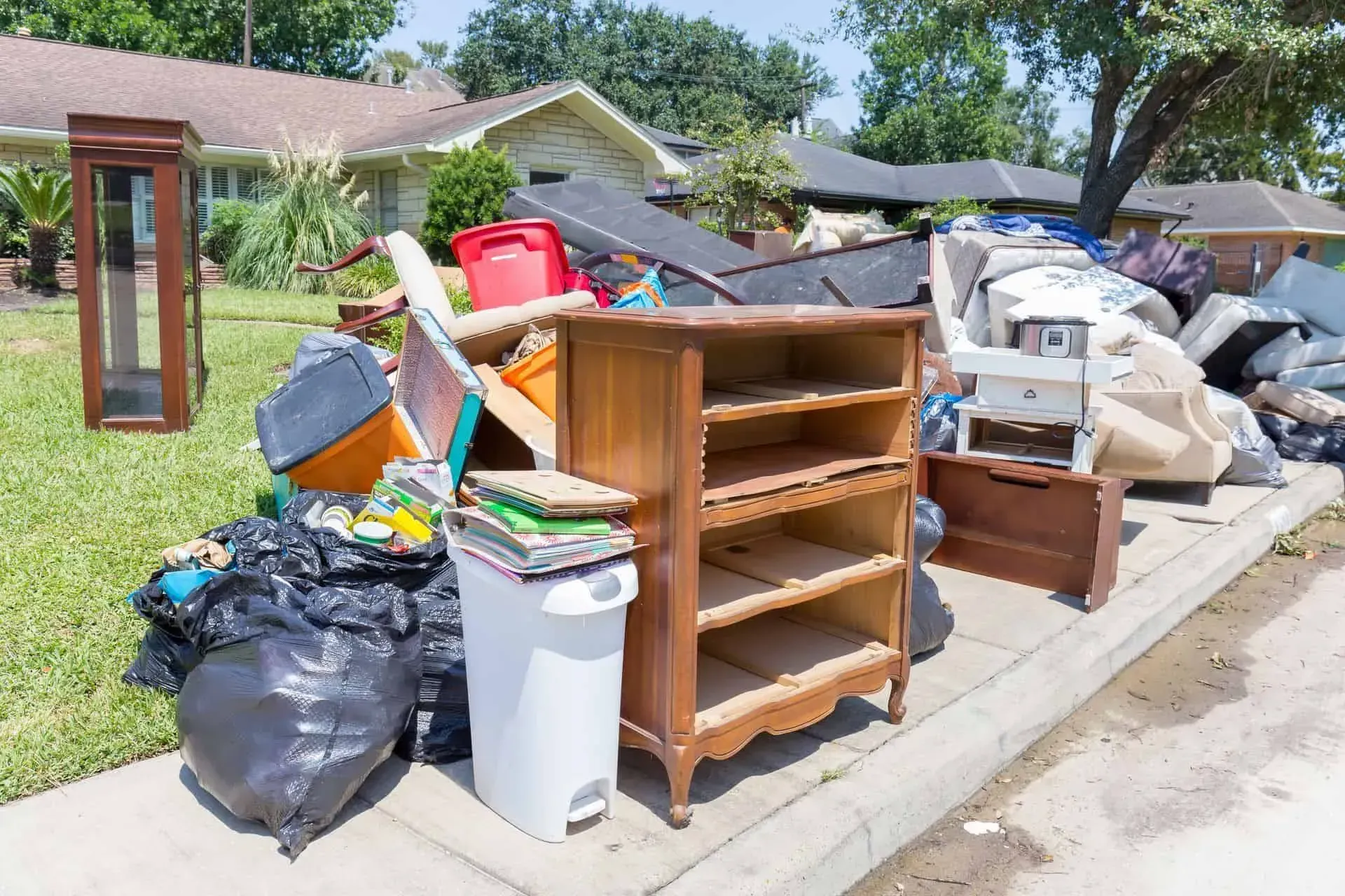 A pile of discarded furniture, trash bags, and household items sits on a suburban sidewalk for curbside pickup.