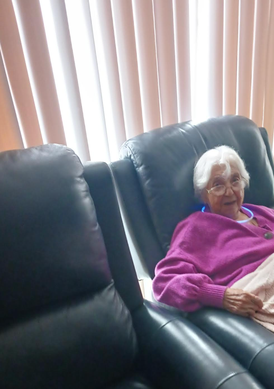 An elderly woman in a purple sweater is sitting on a black leather couch.