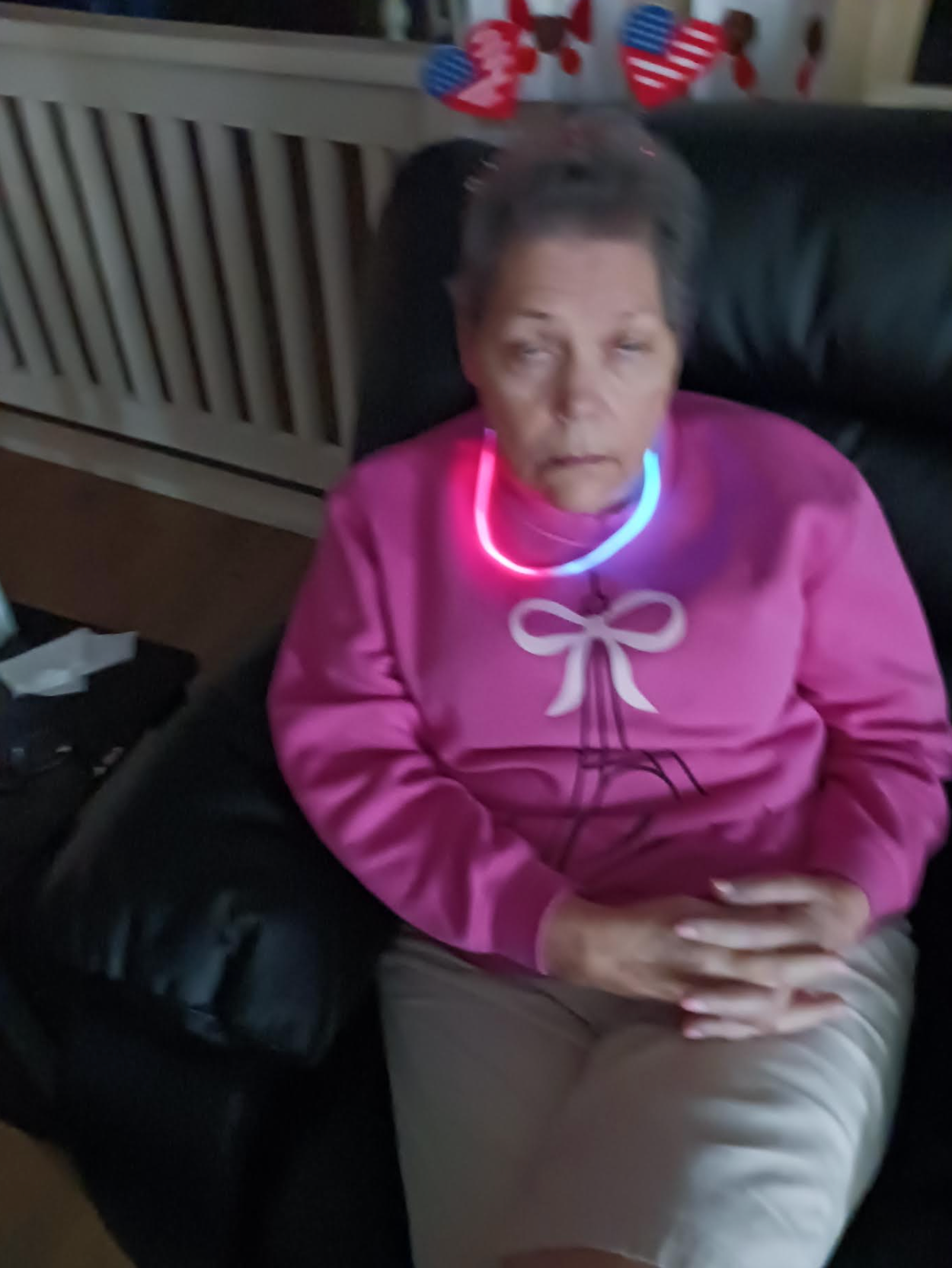An elderly woman is sitting in a chair with a neon necklace around her neck.