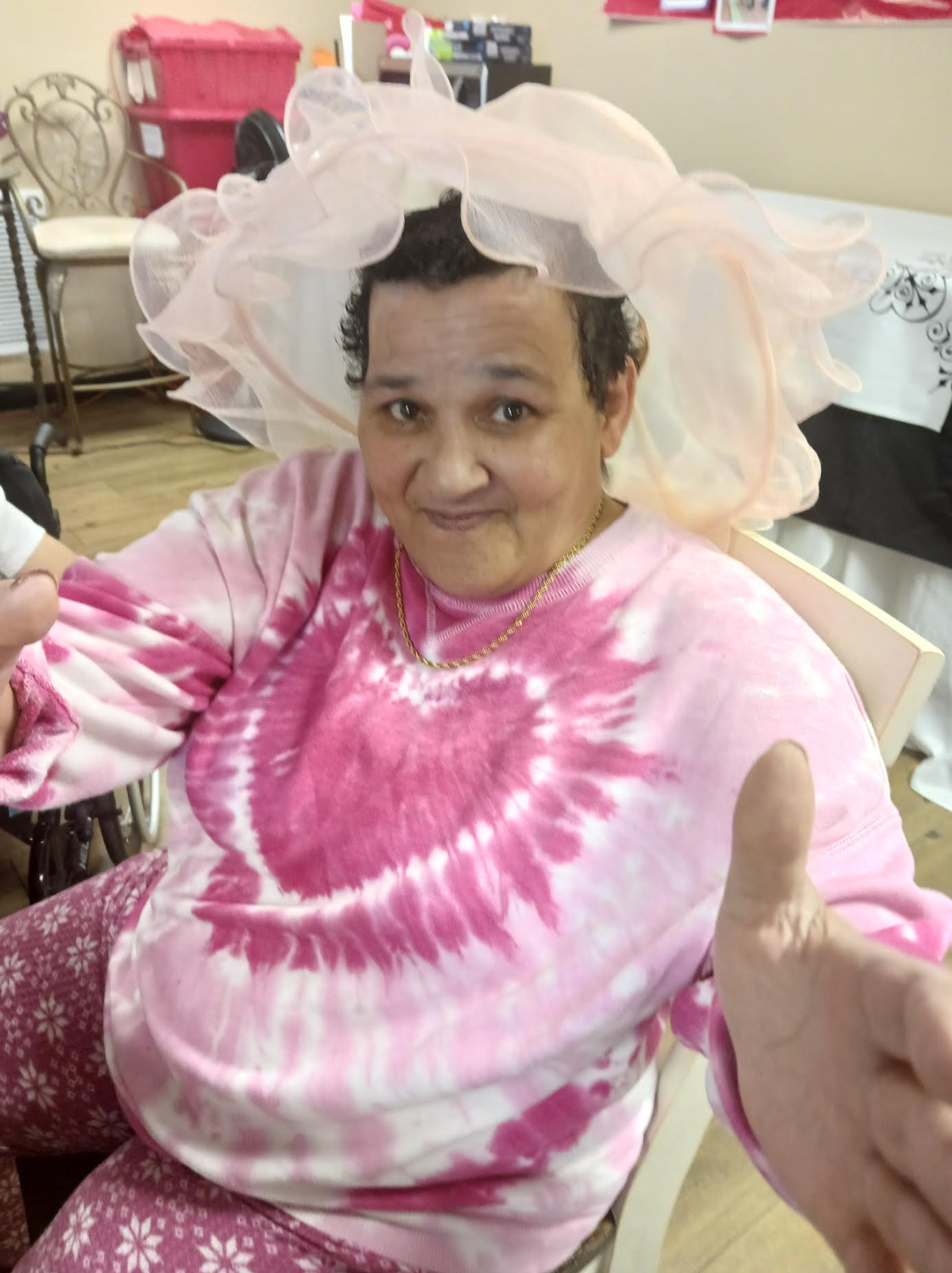 A woman wearing a pink tie dye shirt and a pink hat is giving a thumbs up