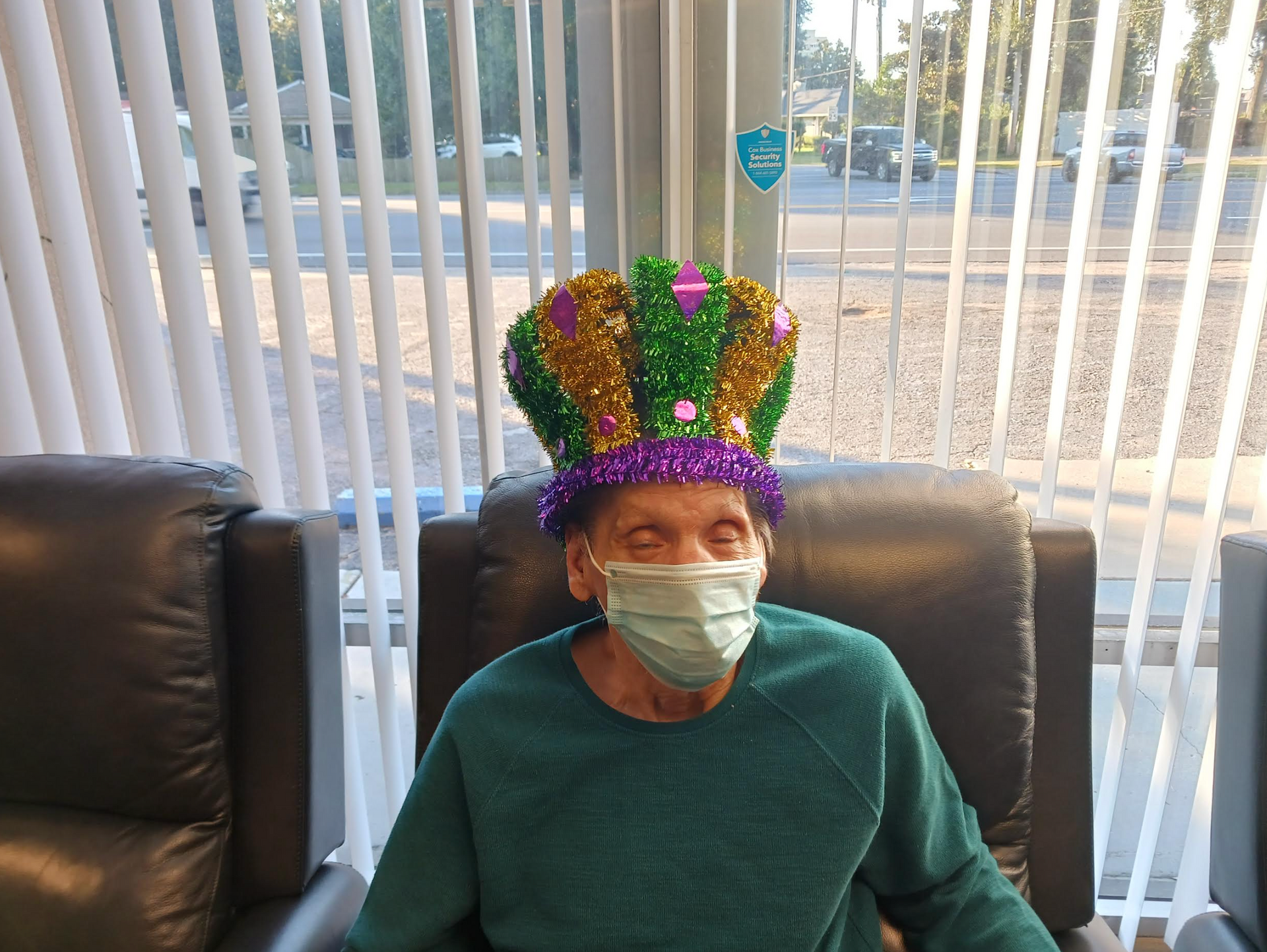 A man wearing a mask and a mardi gras hat is sitting in a chair.