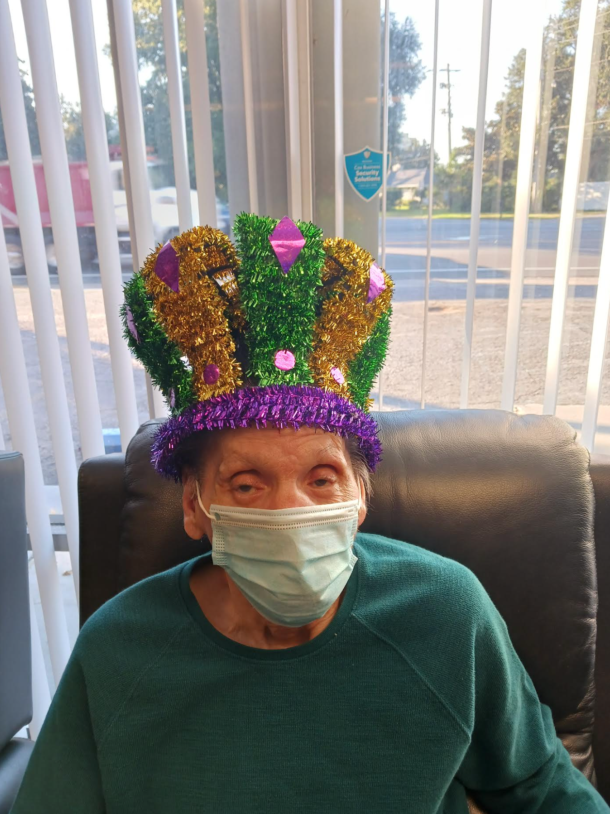 A man wearing a mask and a mardi gras hat is sitting in a chair.