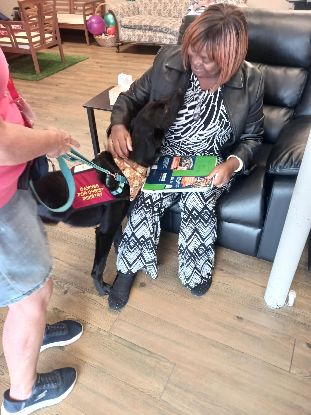 A woman is sitting in a chair reading a book to a dog.