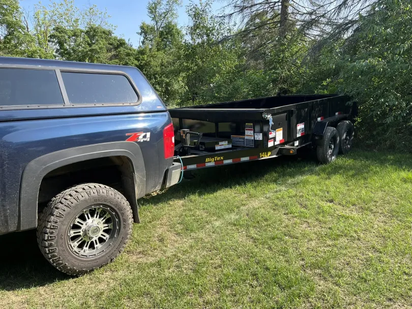 Blue pickup truck towing a black dump trailer on a grassy field, next to trees.