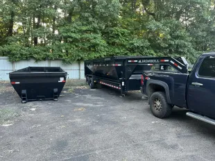 Black truck with a trailer and empty dumpster outdoors, next to a fence and trees.