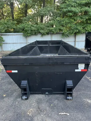 Black dump trailer on wheels with orange and white reflector stripes in front of trees.