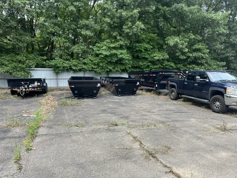 Dumpsters and truck parked on a cracked concrete lot, trees in the background.