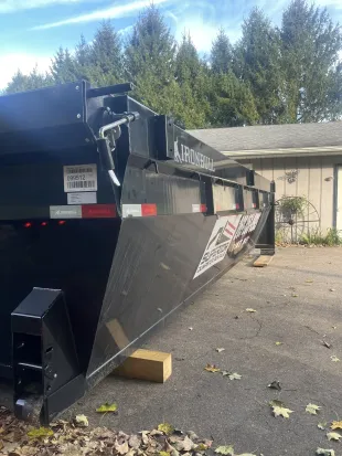 Black dump trailer on wheels with orange and white reflector stripes in front of trees.