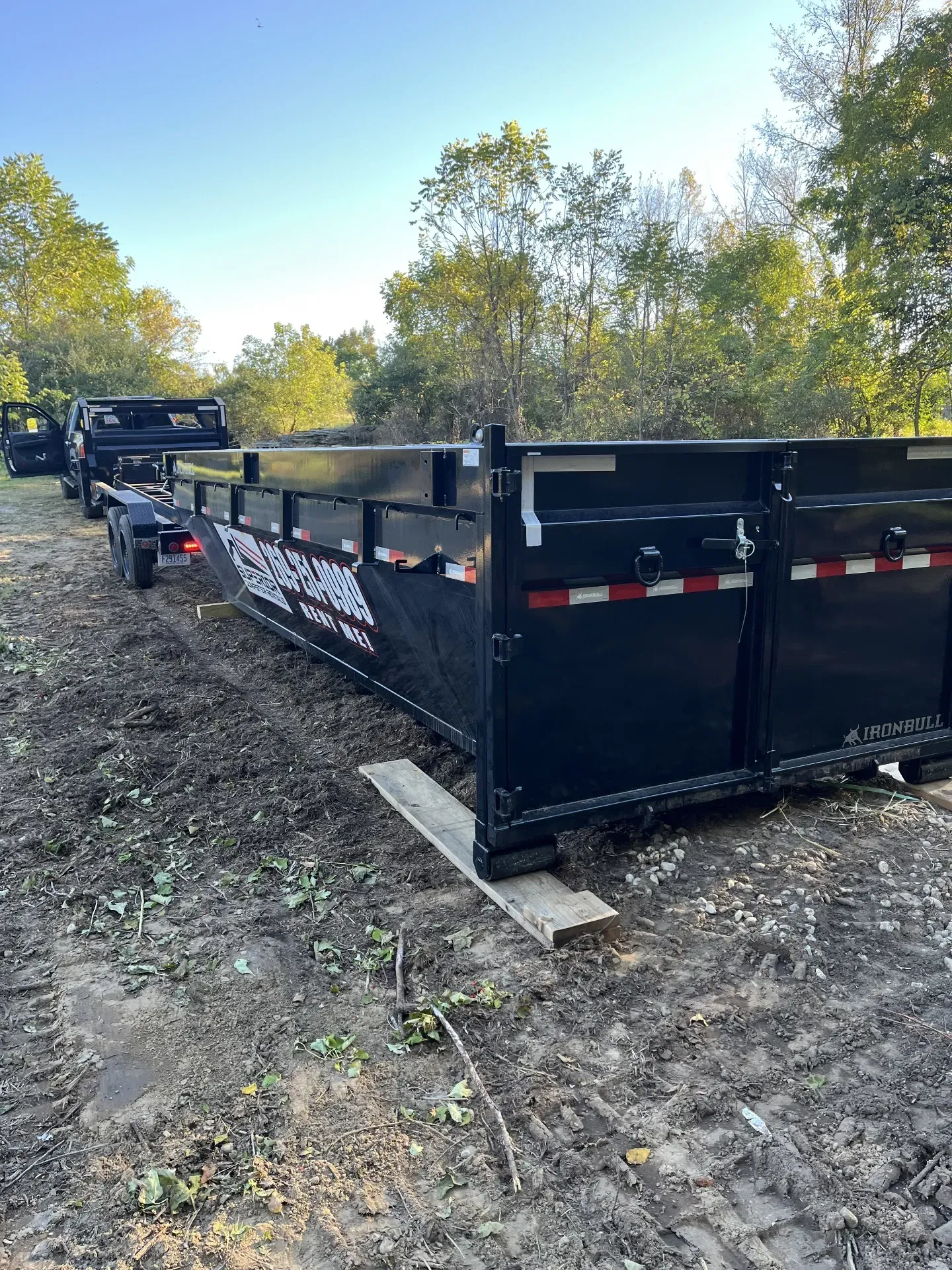 Black truck with a trailer and empty dumpster outdoors, next to a fence and trees.