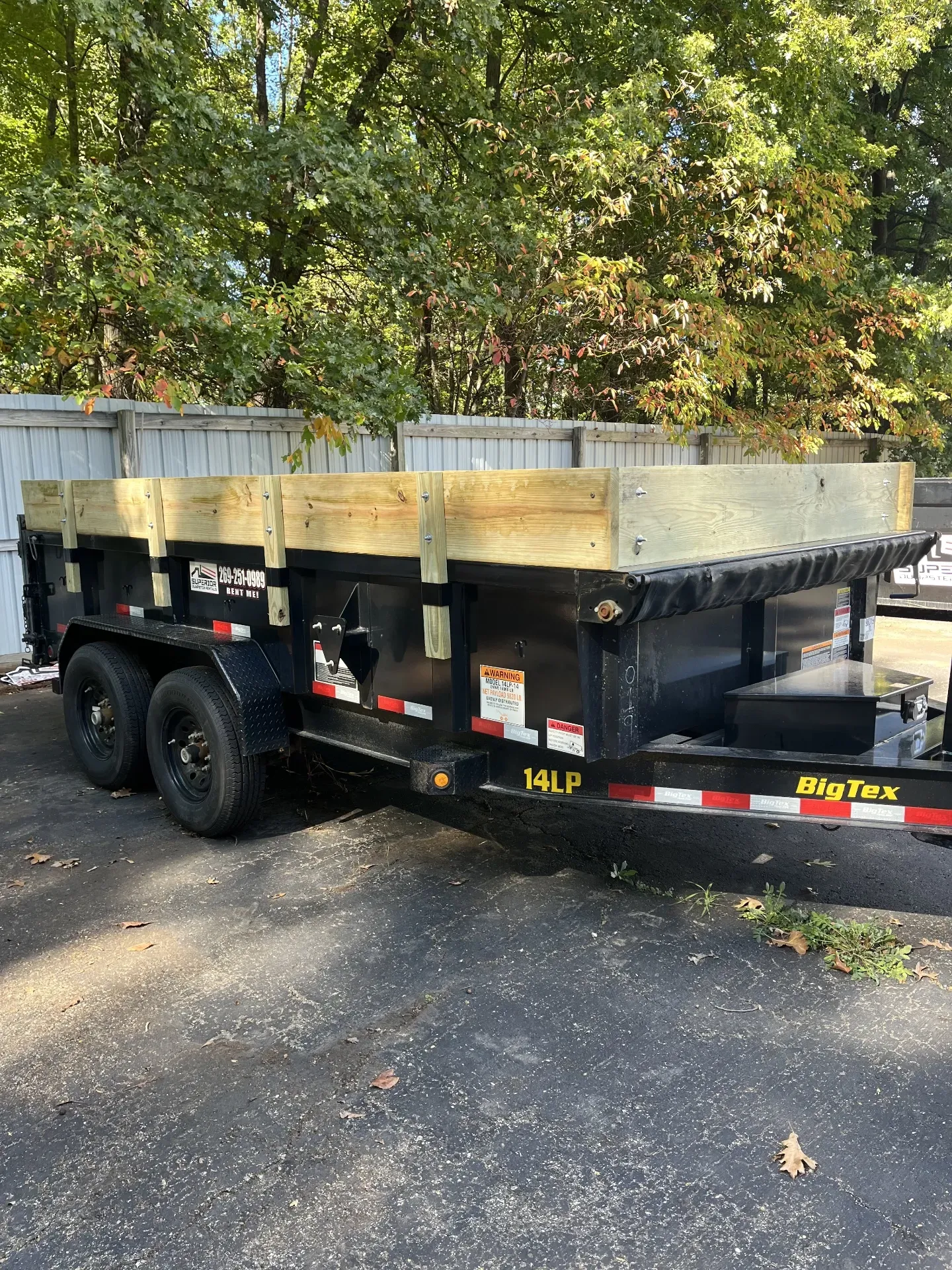 Blue pickup truck towing a black dump trailer on a grassy field, next to trees.