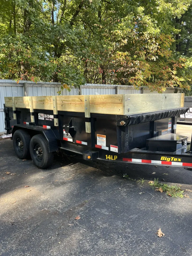 Blue pickup truck towing a black dump trailer on a grassy field, next to trees.