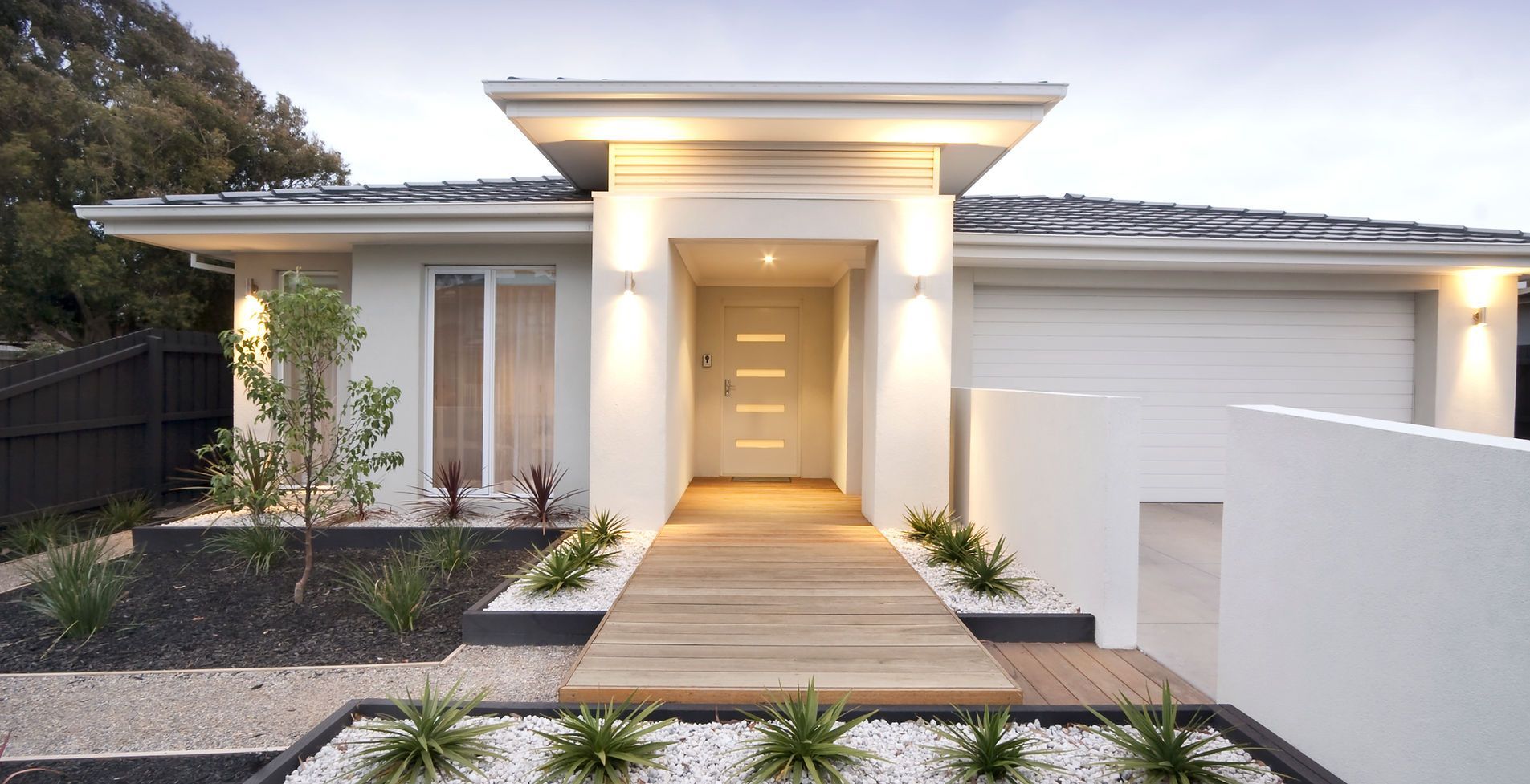 Modern white house with wooden pathway leading to the front door. Landscaping with plants and stone.