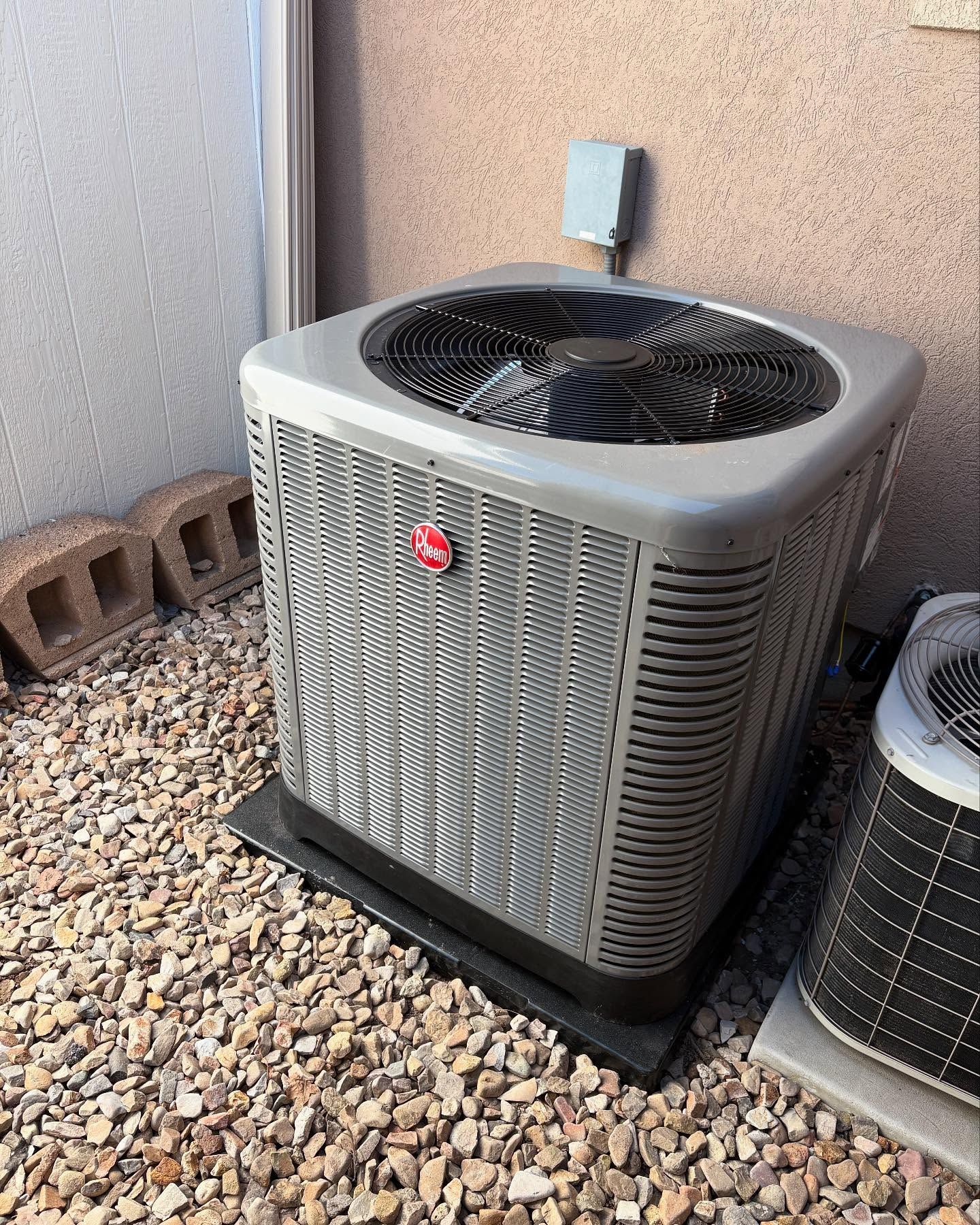 Gray Rheem air conditioner unit on gravel, next to a wall.