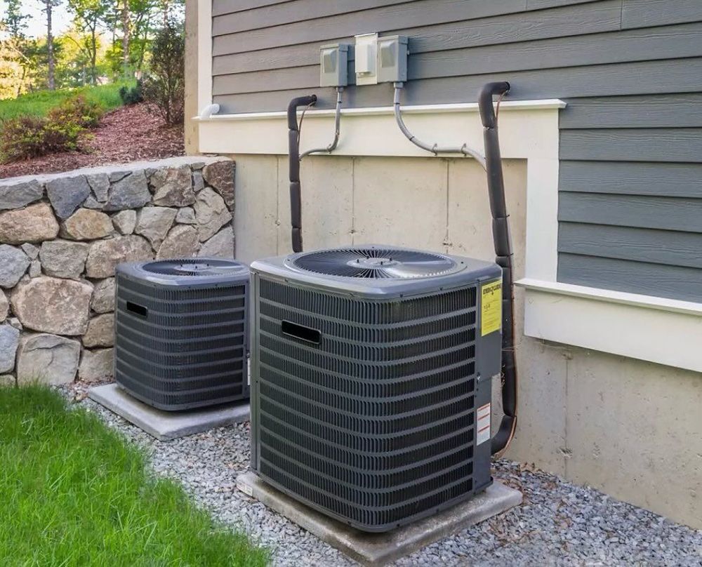 Two outdoor air conditioning units next to a house with gray siding and stone wall. Two outdoor air conditioning units next to a house with gray siding and stone wall.