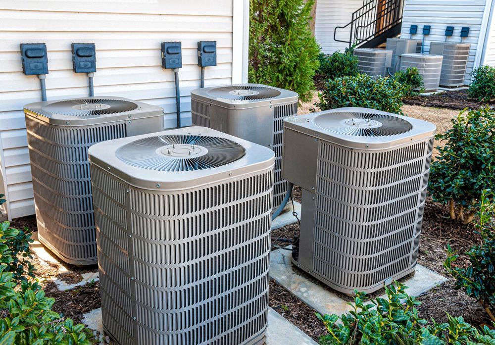 Four gray air conditioning units outside, with electrical boxes above them. Four gray air conditioning units outside, with electrical boxes above them.