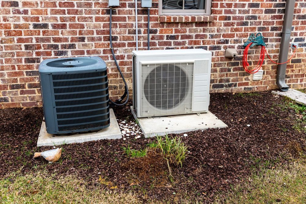 Two outdoor AC units on concrete pads beside a brick wall, with mulch and grass. Two outdoor AC units on concrete pads beside a brick wall, with mulch and grass.