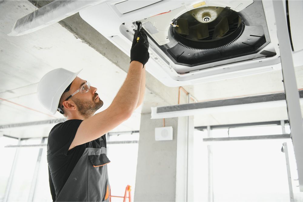 HVAC technician in hard hat repairs ceiling air vent, holding a tool. Interior setting with exposed concrete. HVAC technician in hard hat repairs ceiling air vent, holding a tool. Interior setting with exposed concrete.