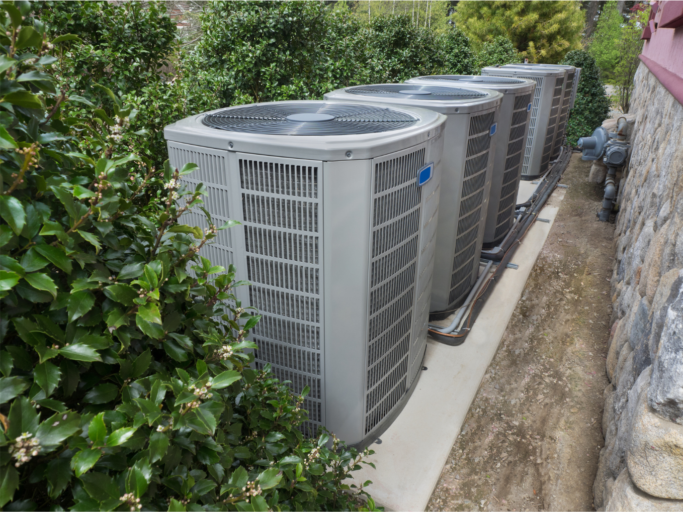 Four air conditioning units aligned, next to bushes, beside a stone wall.
