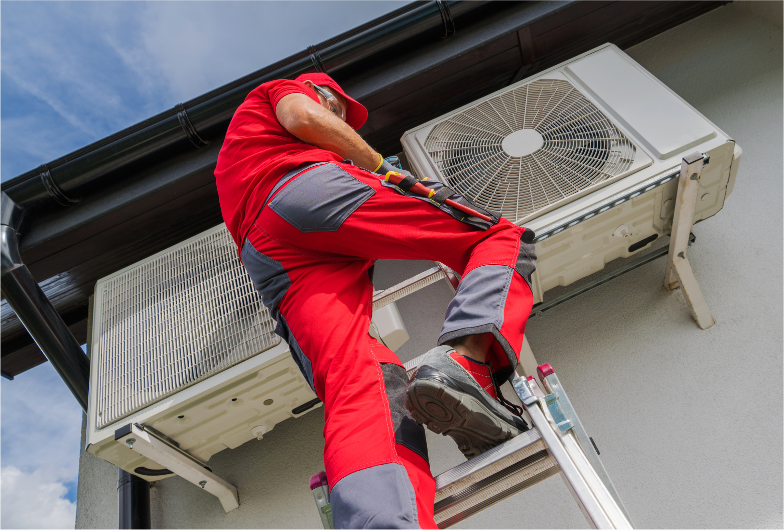 HVAC technician in red overalls on a ladder, working on an outdoor air conditioning unit mounted on a building.