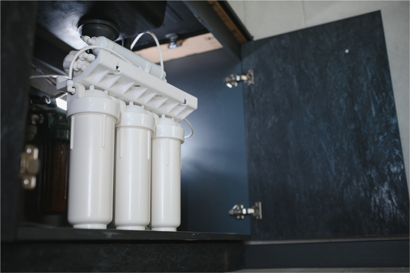 Water filtration system under a dark cabinet. White filters and connecting components. Cabinet door open.