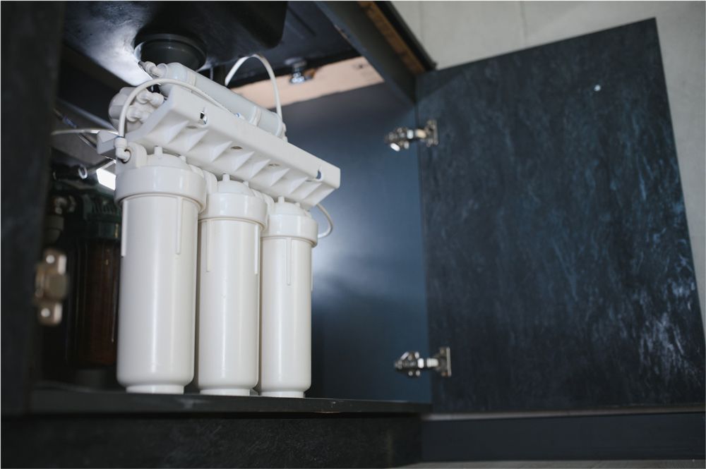Water filtration system under a dark cabinet. White filters and connecting components. Cabinet door open. Water filtration system under a dark cabinet. White filters and connecting components. Cabinet door open.