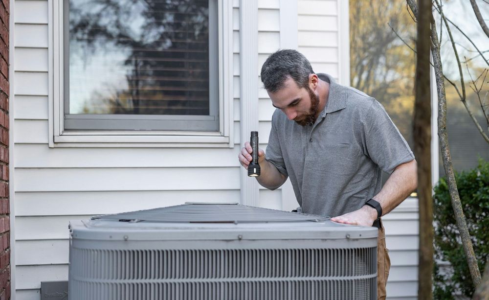Man inspecting an outdoor AC unit with a flashlight next to a house.