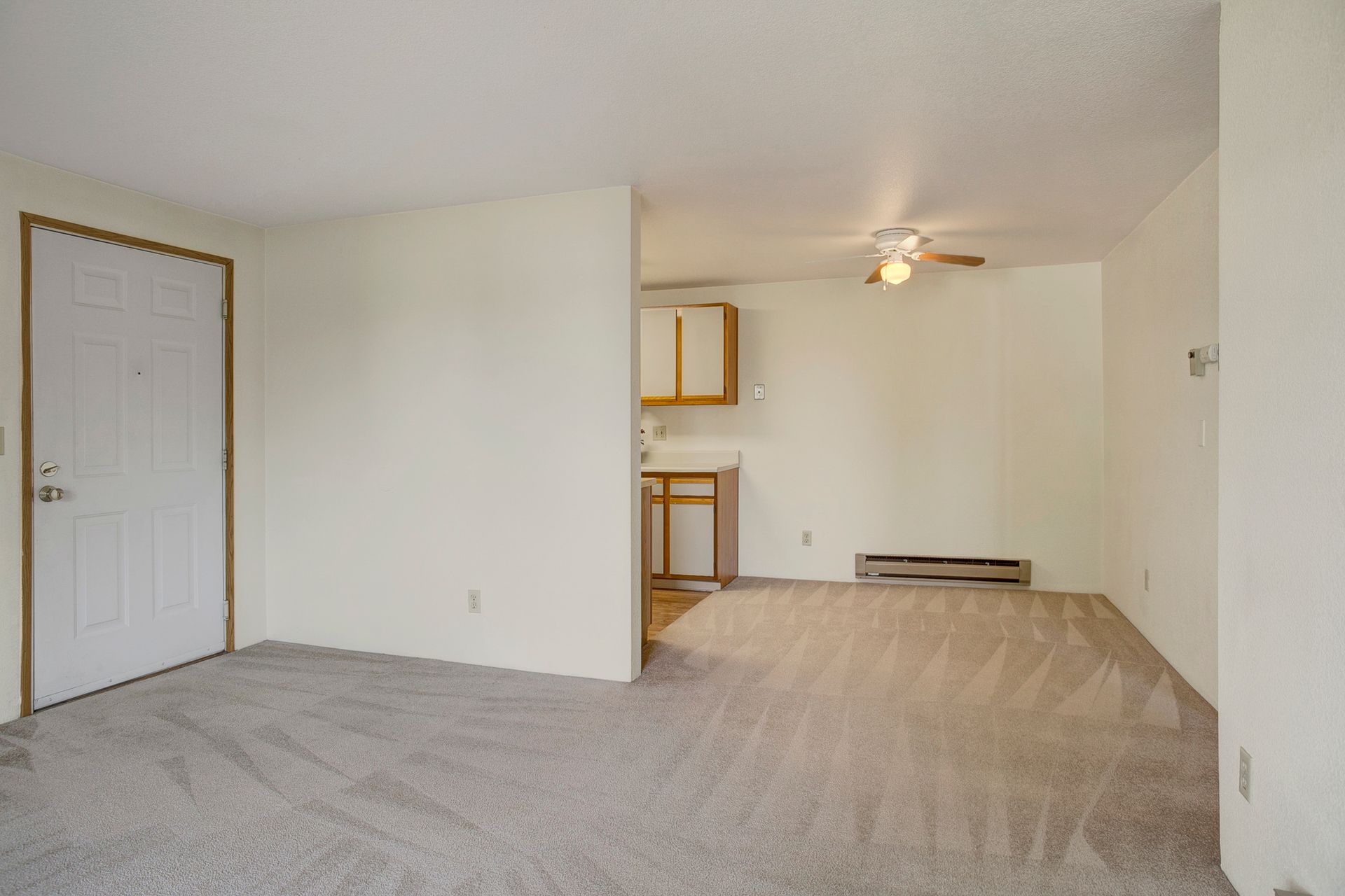 An empty living room with a ceiling fan and a kitchen in the background