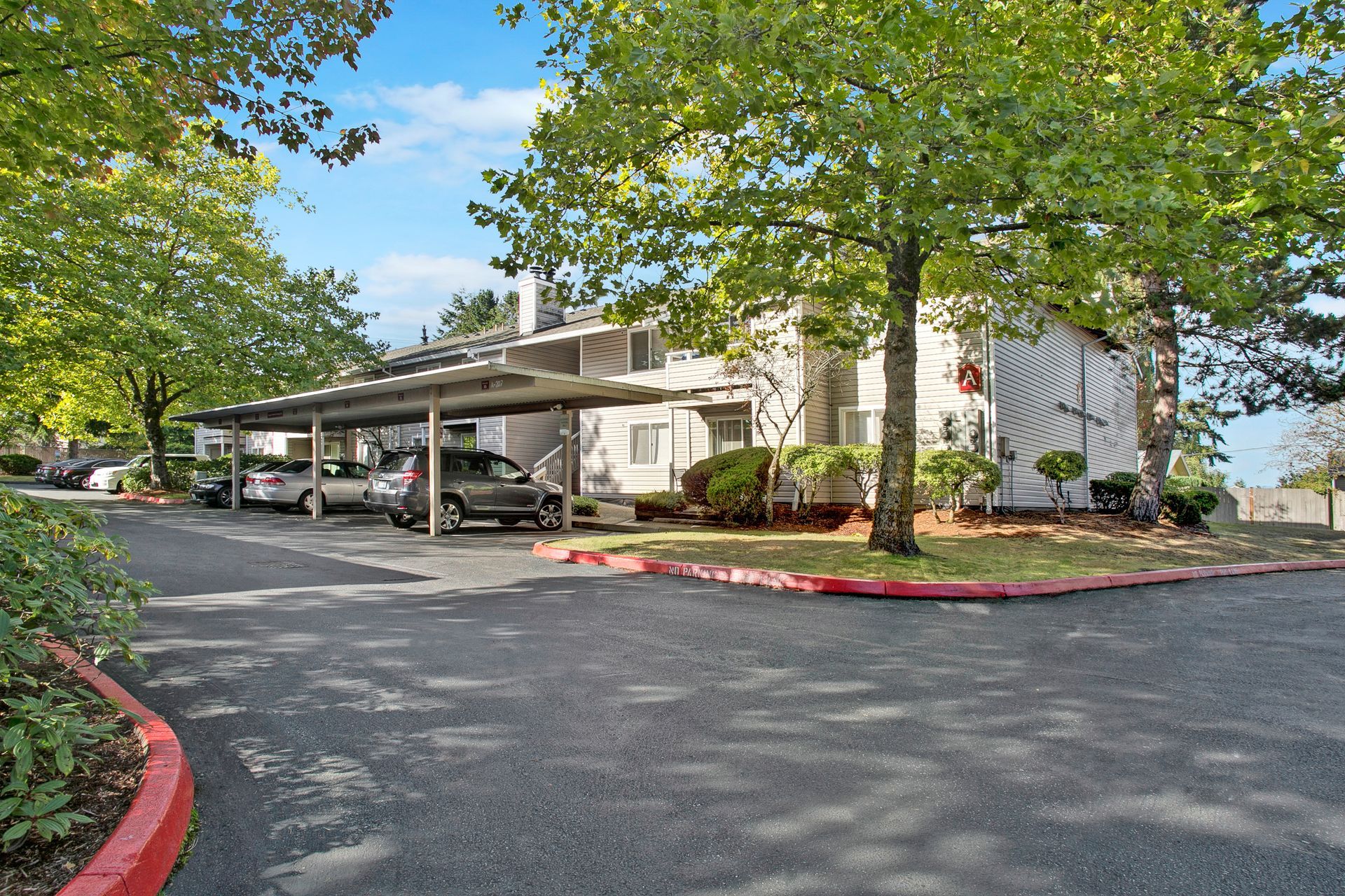 A row of cars are parked under a covered parking lot in front of a building.