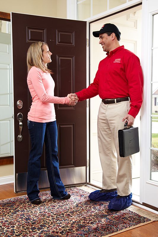 Woman shaking hands with a man at a doorway; man wears a red shirt and blue shoe covers.