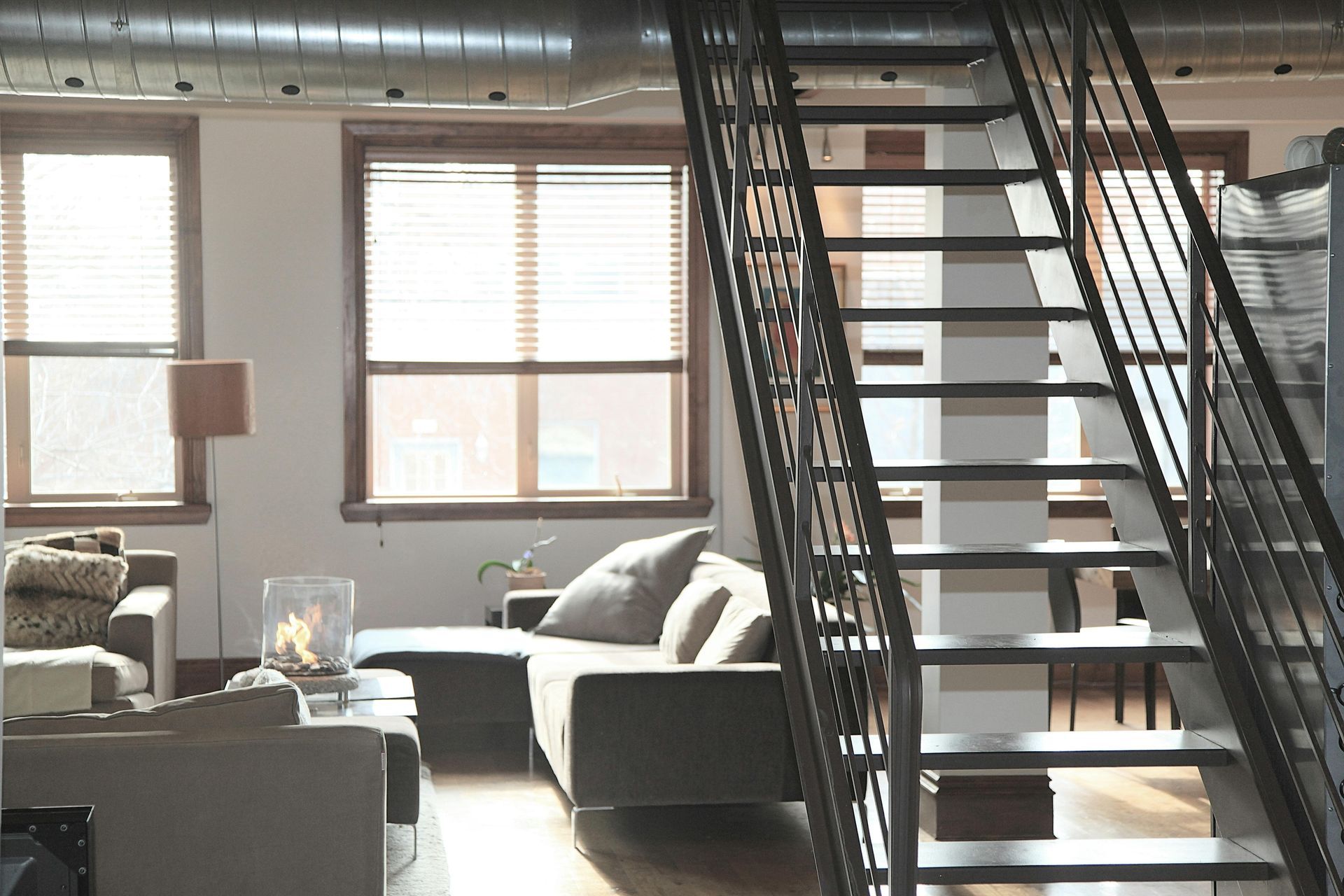 Living room with stairs, couch, windows, and exposed ductwork. Sunlight streams in.