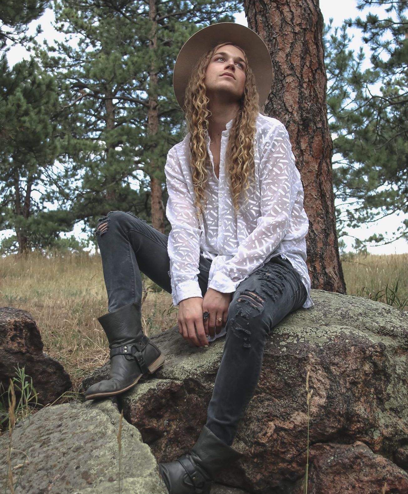 A person with long hair is sitting on a rock under a tree for a photo shoot.