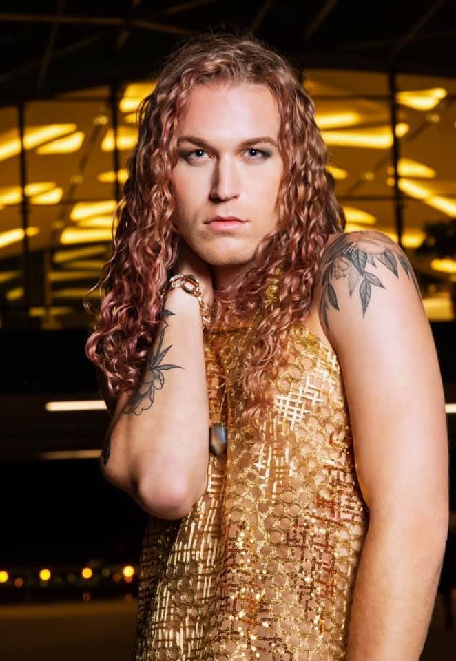 A man with long curly hair and a tattoo on his arm is wearing a gold dress.