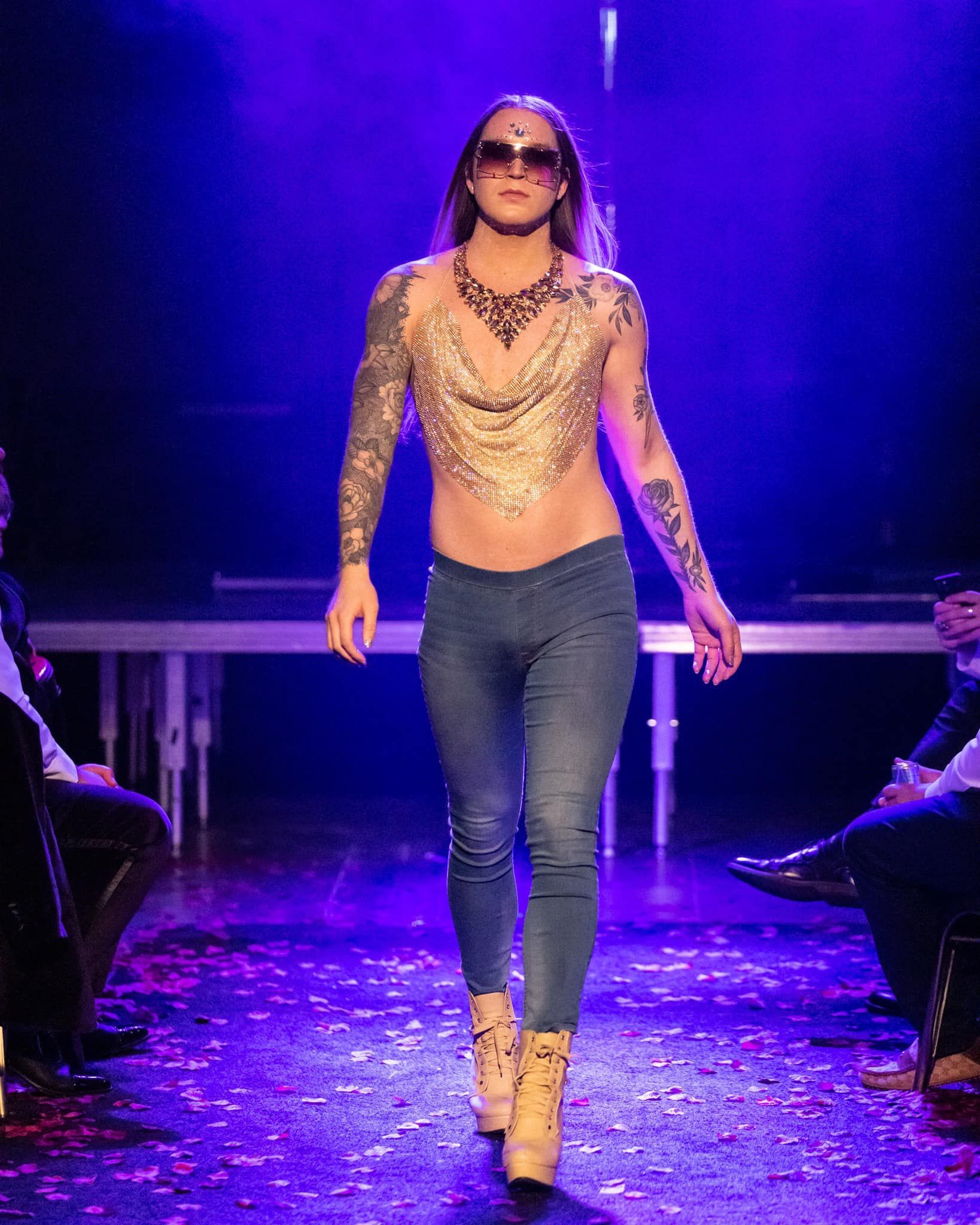 A man is walking down a runway wearing a necklace and sunglasses.
